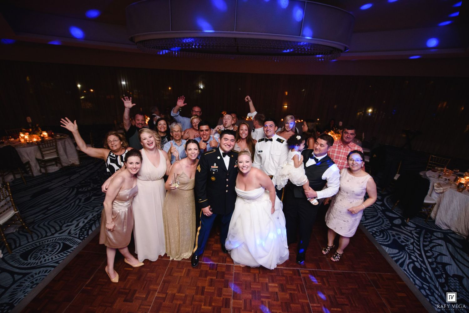 Wedding party group photo on dance floor with bride and groom in center, guests celebrating at elegant reception.