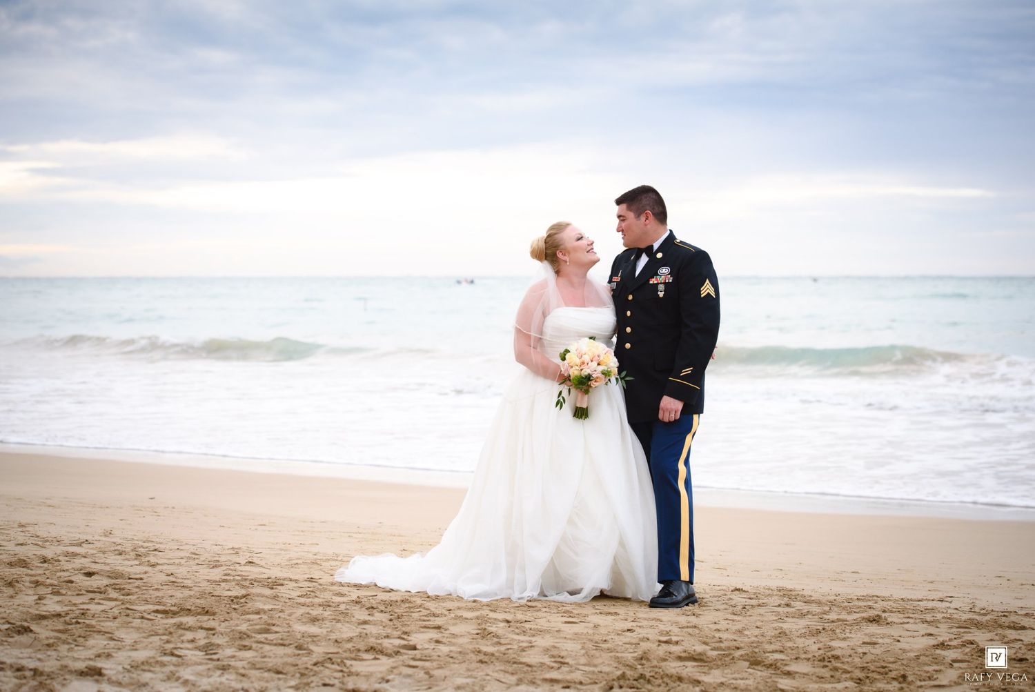 Bride and groom in military dress uniform share a romantic moment on a sandy beach with ocean waves behind them.