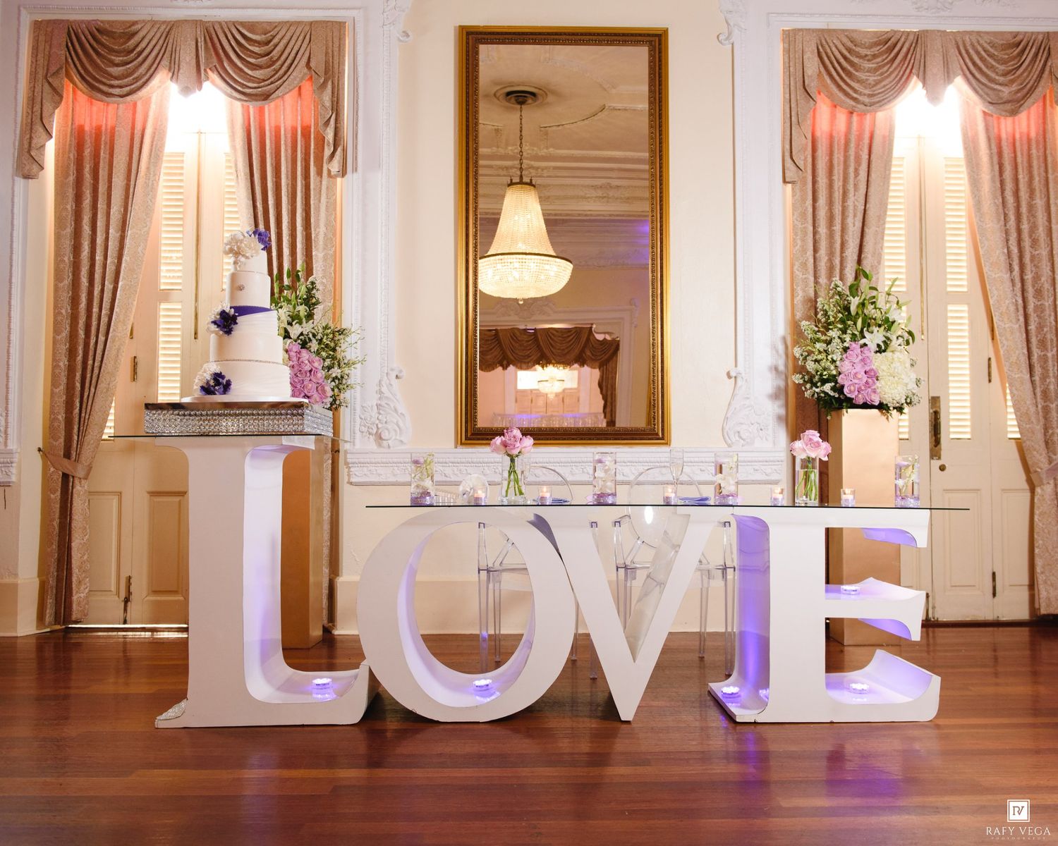 Elegant wedding venue with large illuminated LOVE letters on hardwood floor before a fireplace with floral arrangements.