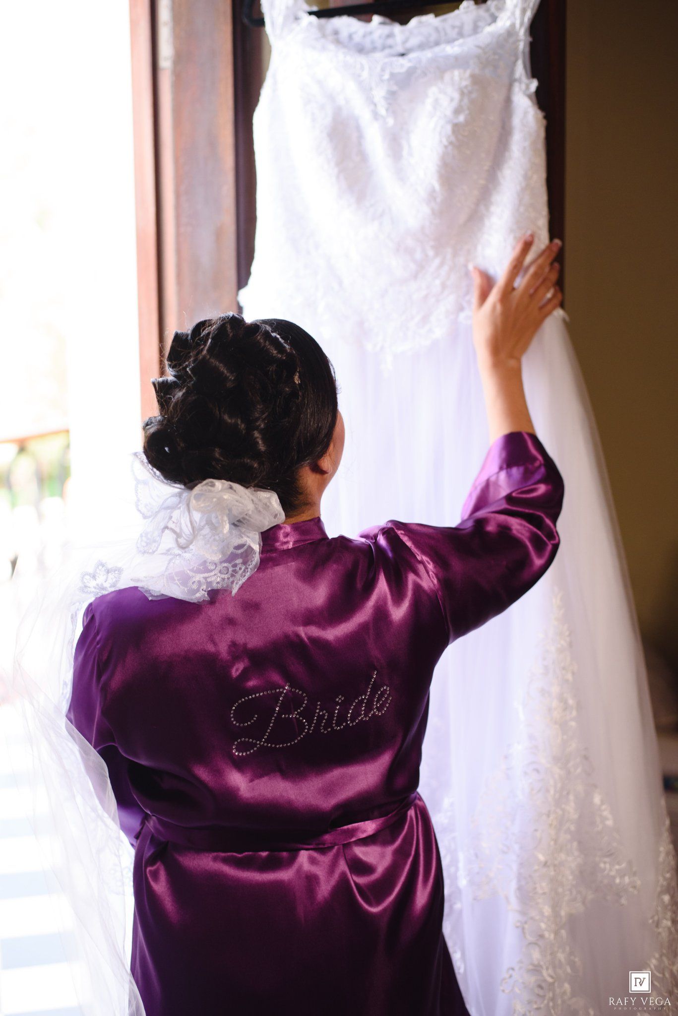 Bride in purple satin robe touching her white lace wedding dress hanging by a window on her wedding day.