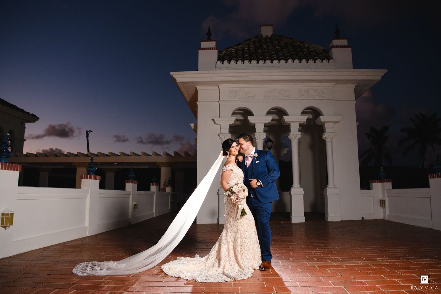 Bride and groom share romantic kiss on rooftop terrace at dusk, her long veil flowing behind them.