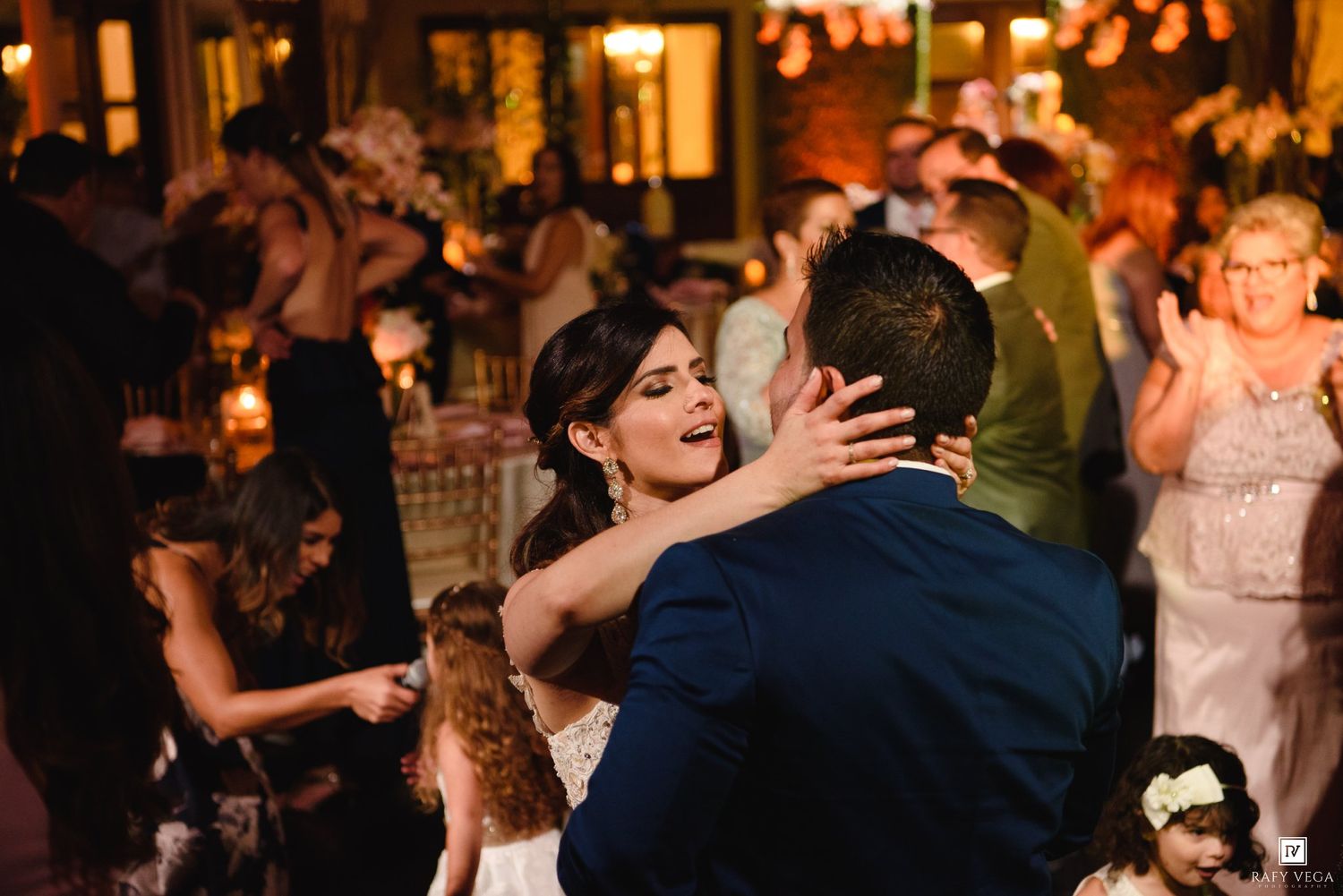 Bride and groom share their first dance at a warmly lit wedding reception surrounded by celebrating guests.