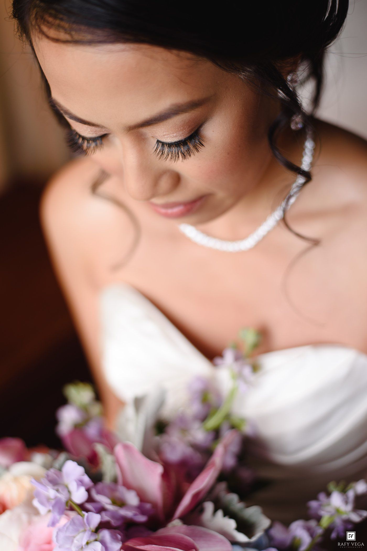 Bride in white strapless gown holding purple and pink floral bouquet, wearing pearl necklace with elegant bridal makeup.