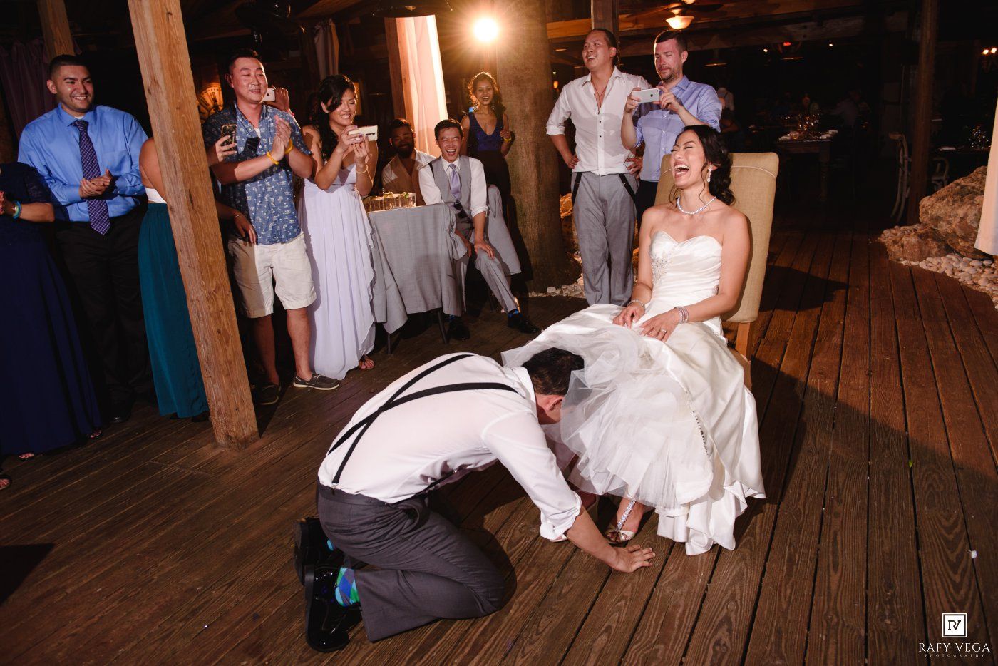 Groom removing bride's garter while guests cheer at a rustic wedding reception.