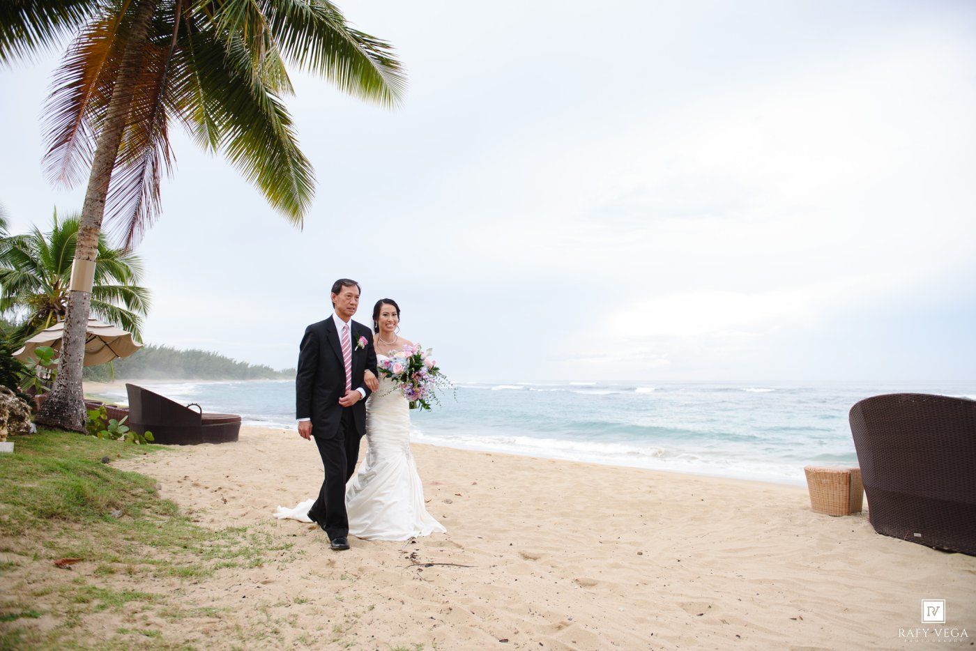 Bride and groom walking on tropical beach with palm trees and ocean backdrop.