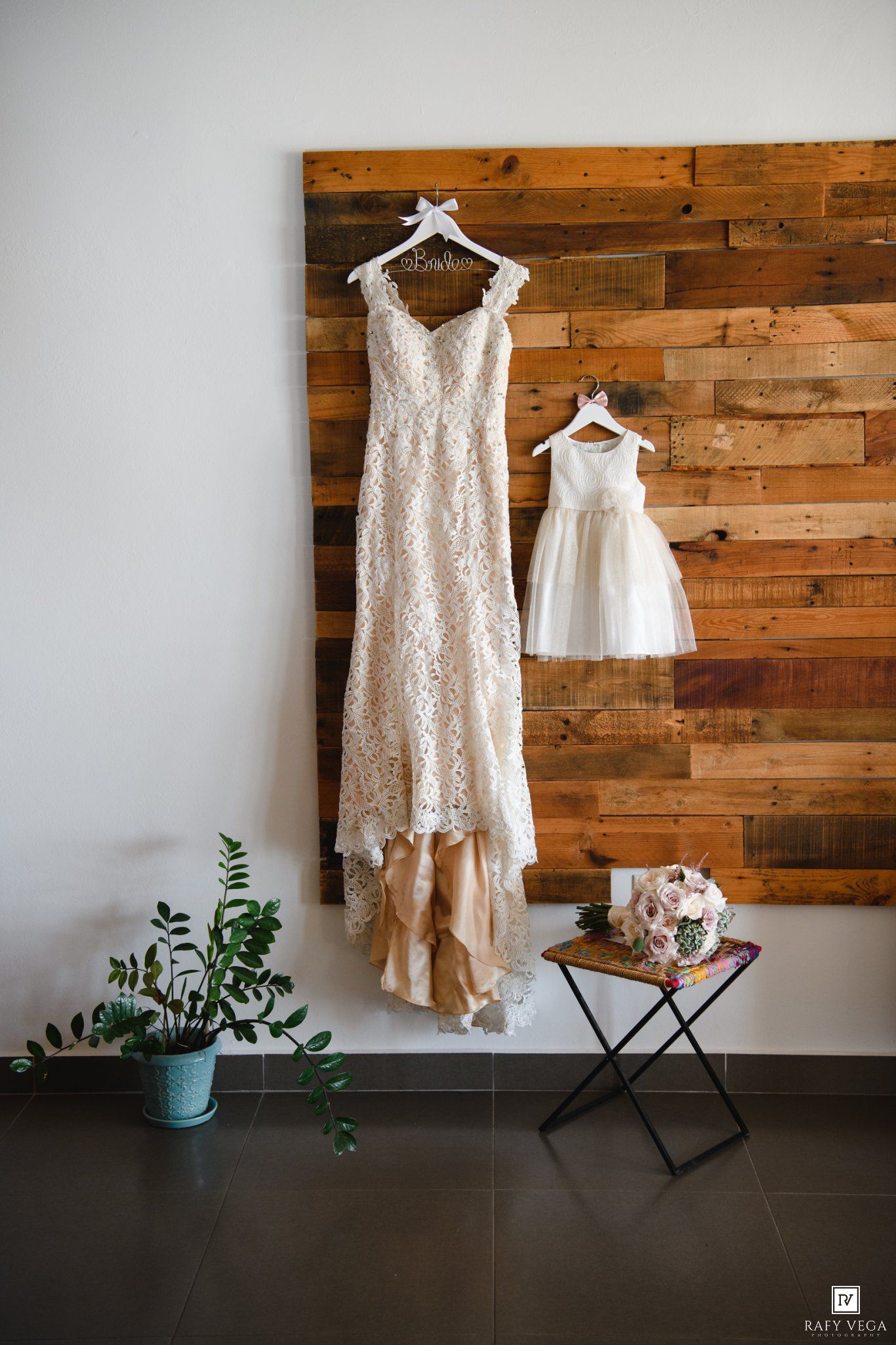 Elegant lace wedding gown and flower girl dress hanging on rustic wood plank wall with bridal bouquet on side table.
