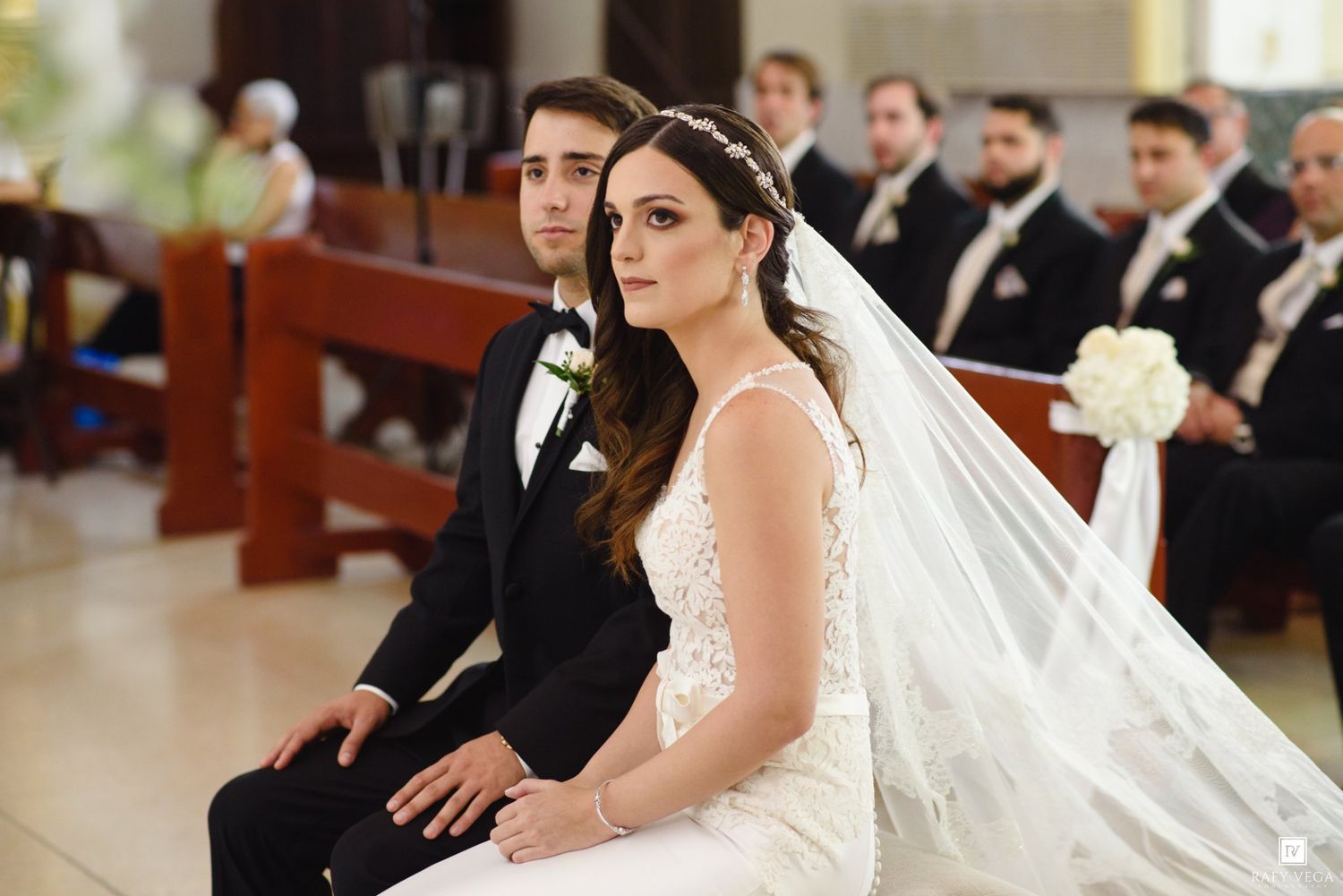 Bride and groom seated together during wedding ceremony in church, bride wearing lace gown and veil.