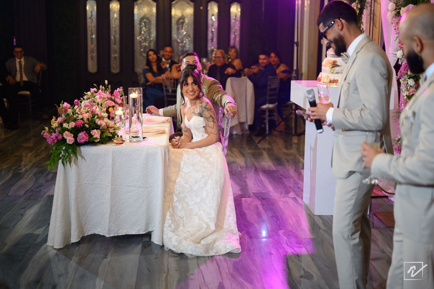 Bride in a white gown kneels beside a table with pink flowers at a wedding reception while guests and two men in suits look on.