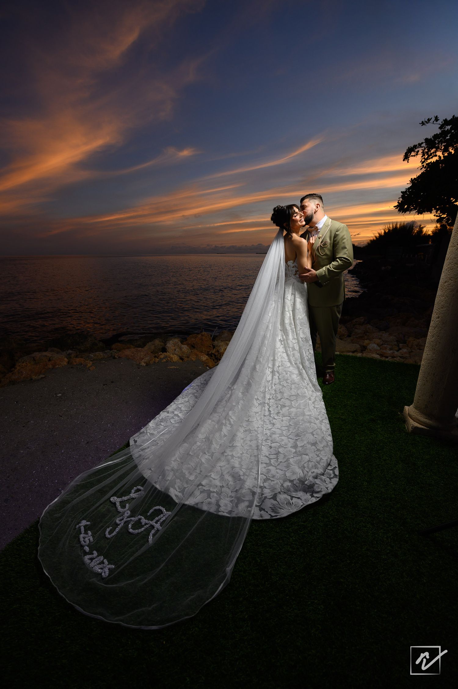 Bride in a lace gown with a long embroidered veil kisses her groom on a seaside lawn at sunset under a colorful sky.