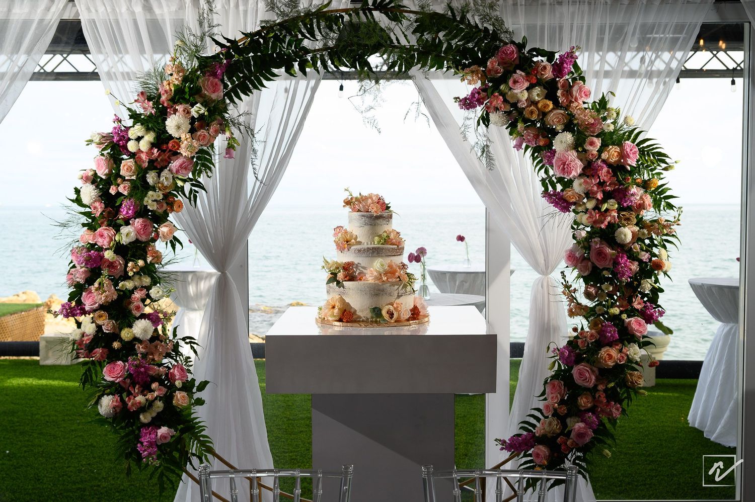 Three-tier wedding cake on a white table beneath a floral arch of pink and white roses, with sheer drapes and an ocean view behind.