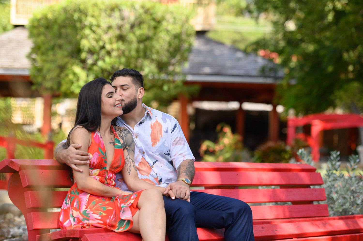 Couple sharing a romantic kiss on a red bench in a lush garden setting, woman in floral dress, man in blue shirt.