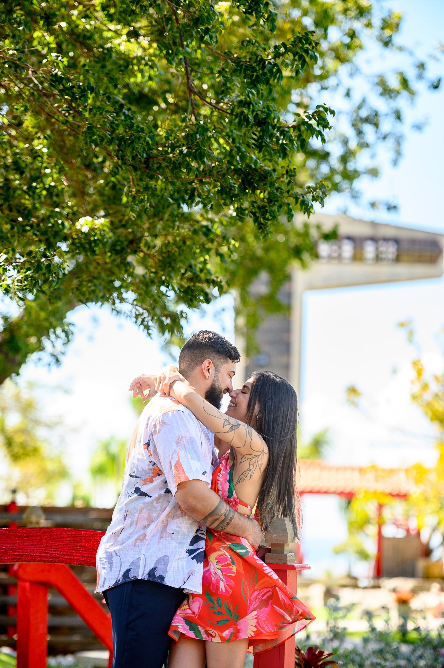 Couple embracing and kissing under a lush green tree near a red bench in a tropical outdoor setting.