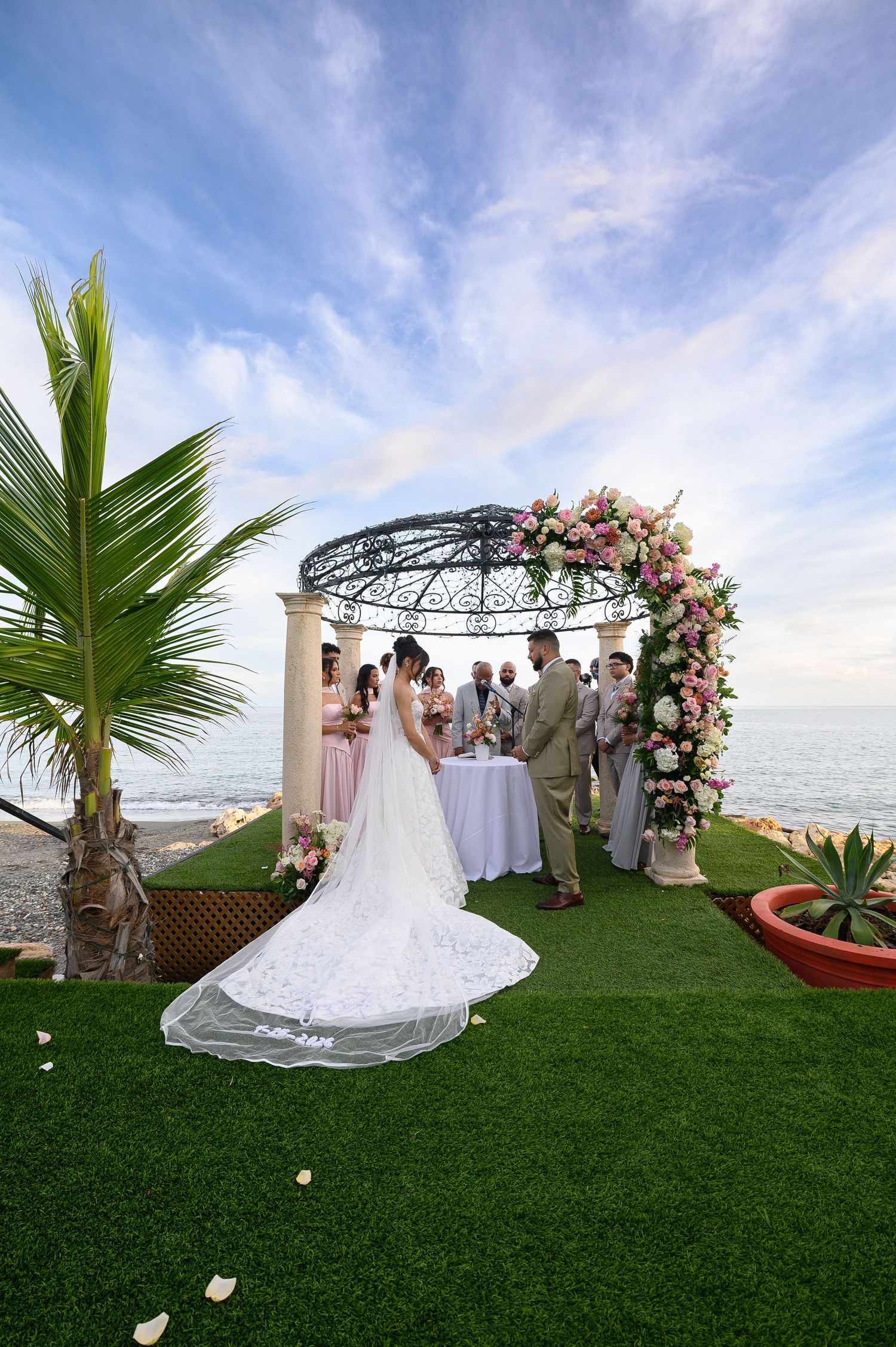 Romantic beachside wedding ceremony at Ocean View Ponce under a floral arch gazebo with bride in lace gown, bridesmaids, and ocean backdrop.