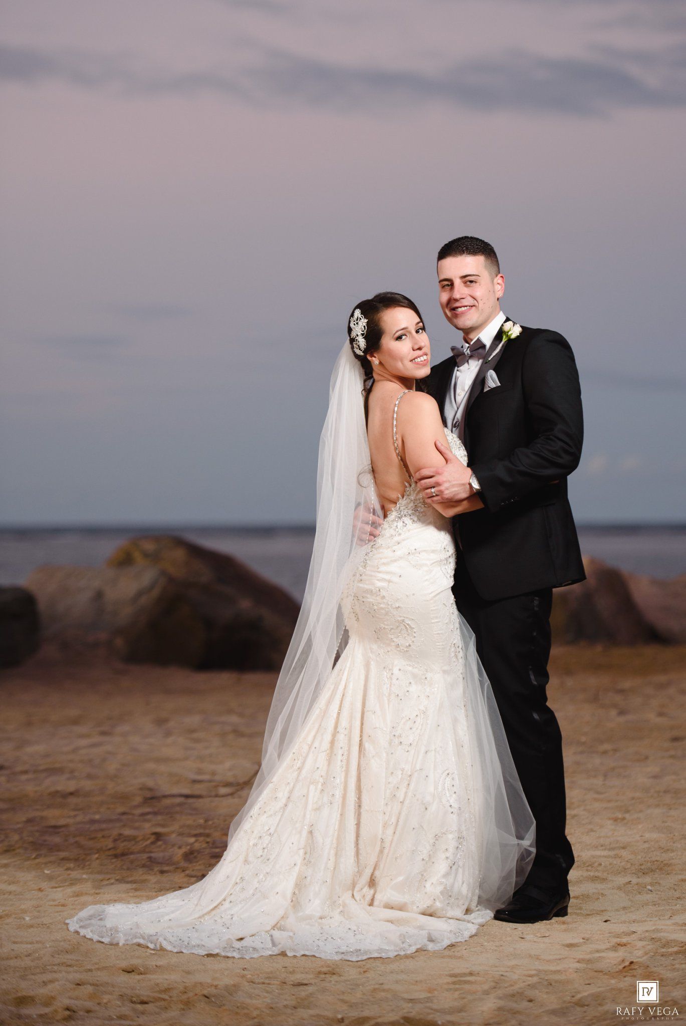 Bride in lace mermaid gown and veil poses with groom in black tuxedo on sandy beach at sunset.