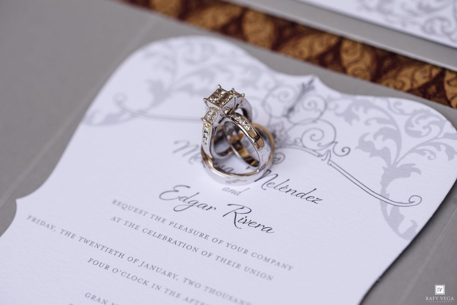 Elegant wedding invitation for Edgar and Milindee Rivera with diamond engagement ring and wedding bands resting on top.