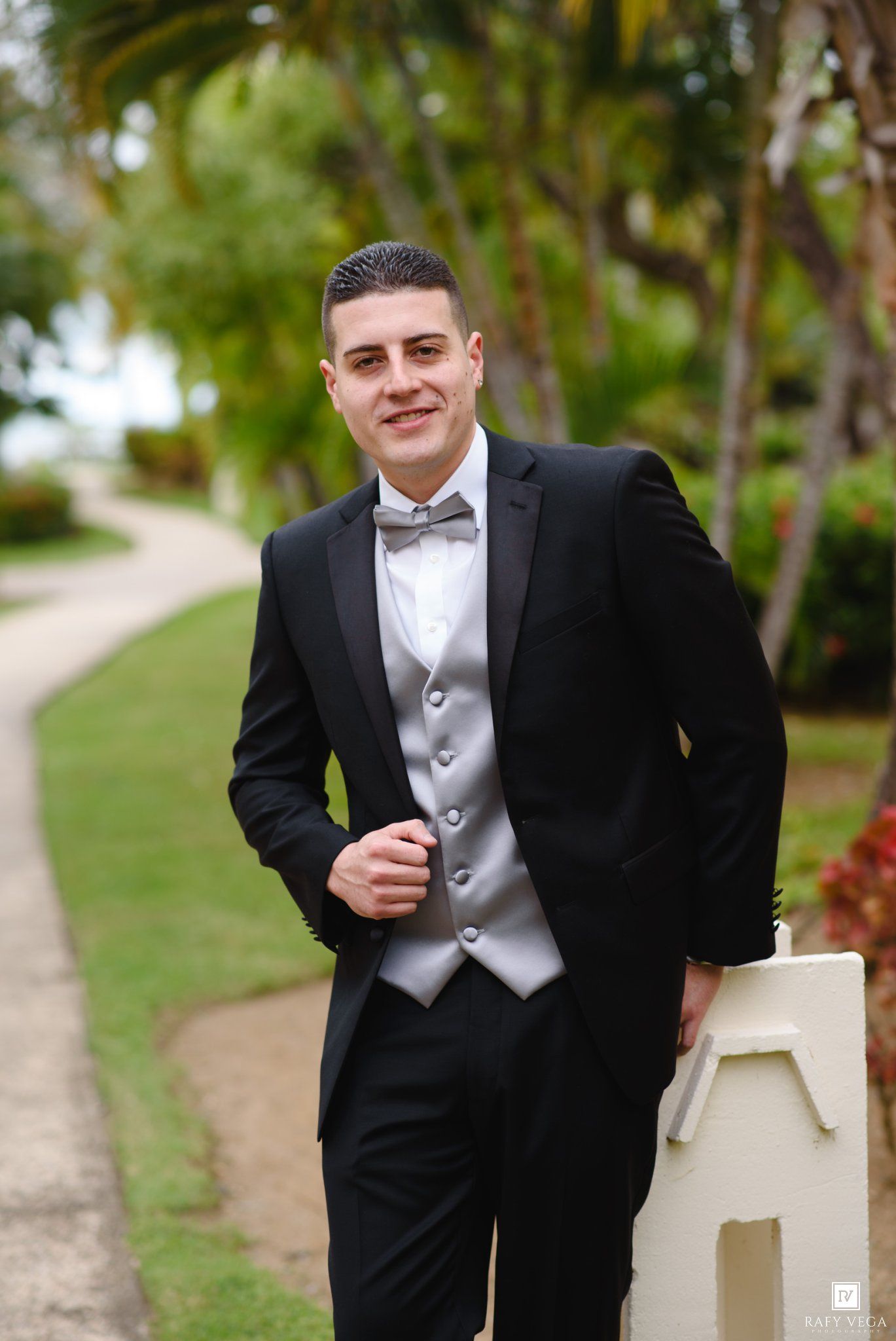 Smiling groom in black tuxedo with silver vest and bow tie posing outdoors among tropical palm trees.