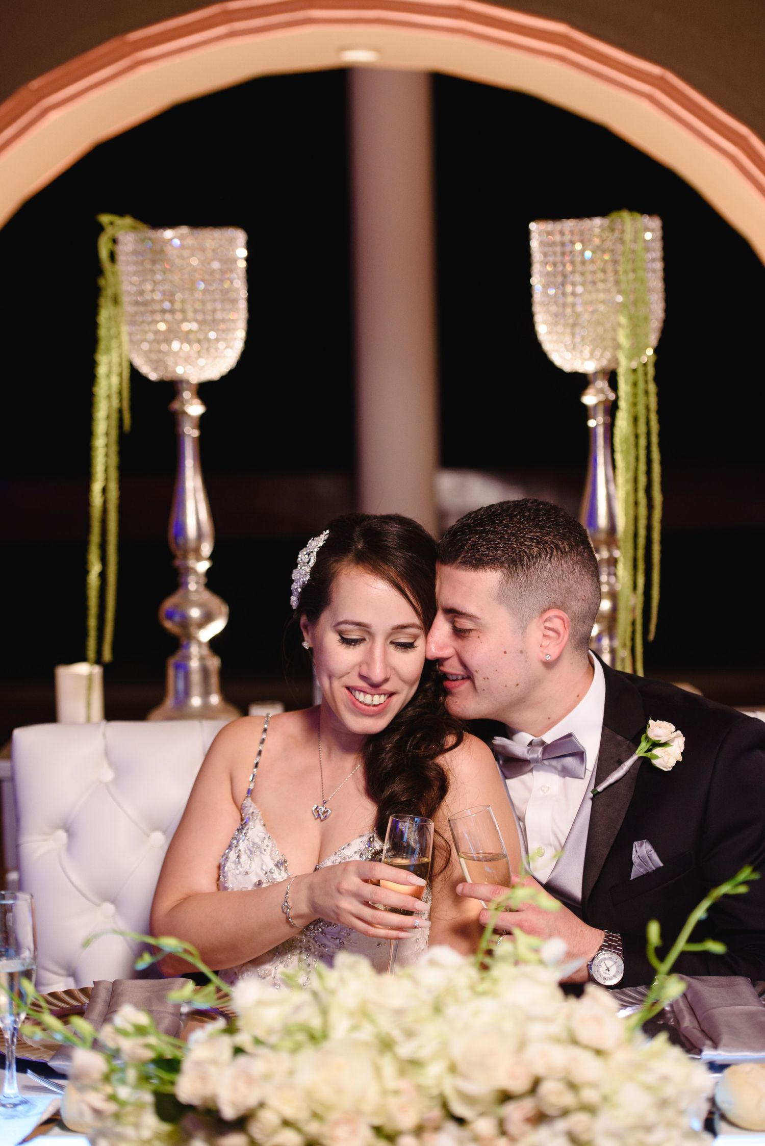 Bride and groom share a romantic toast at their wedding reception, surrounded by elegant crystal centerpieces and white floral arrangements.