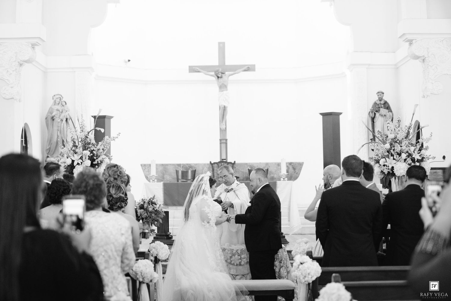 Black and white wedding ceremony photo inside a church with bride and groom at the altar beneath a crucifix.