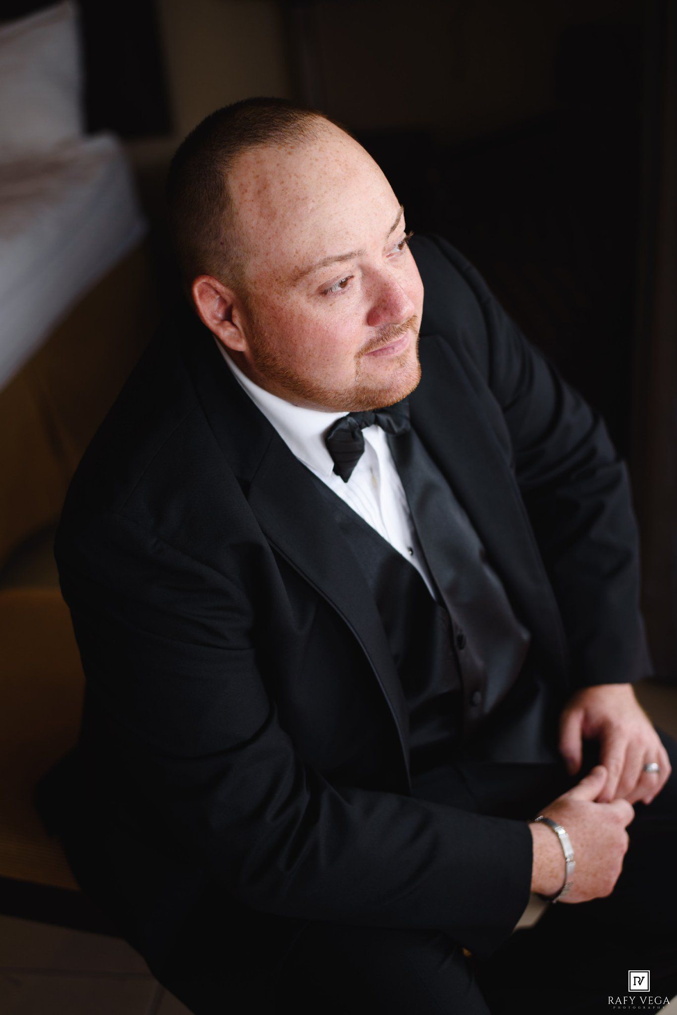 A well-dressed groom in a black tuxedo with bow tie sits thoughtfully, looking away in a dimly lit elegant setting.