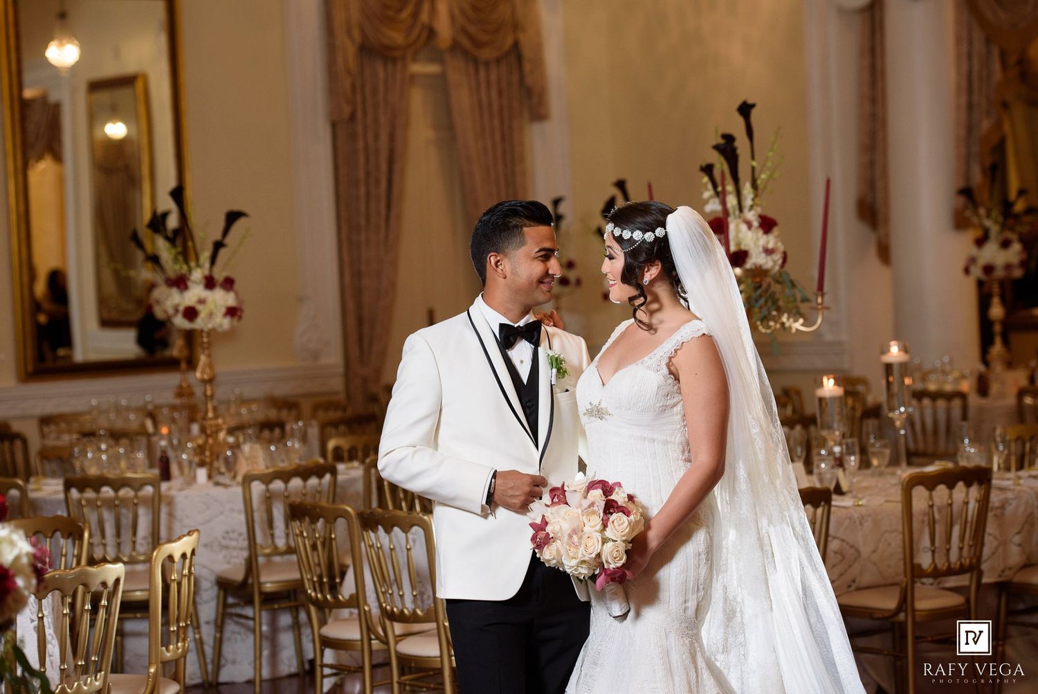 Bride and groom share a romantic moment in a grand ballroom with gold chairs and floral wedding decor.