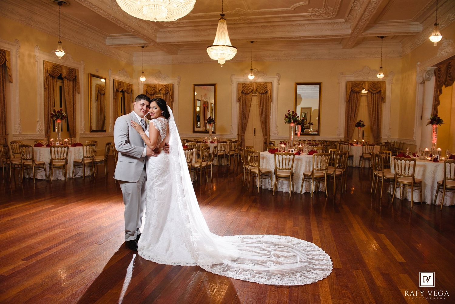Bride and groom embrace in elegant wedding venue with chandeliers and gold chairs.