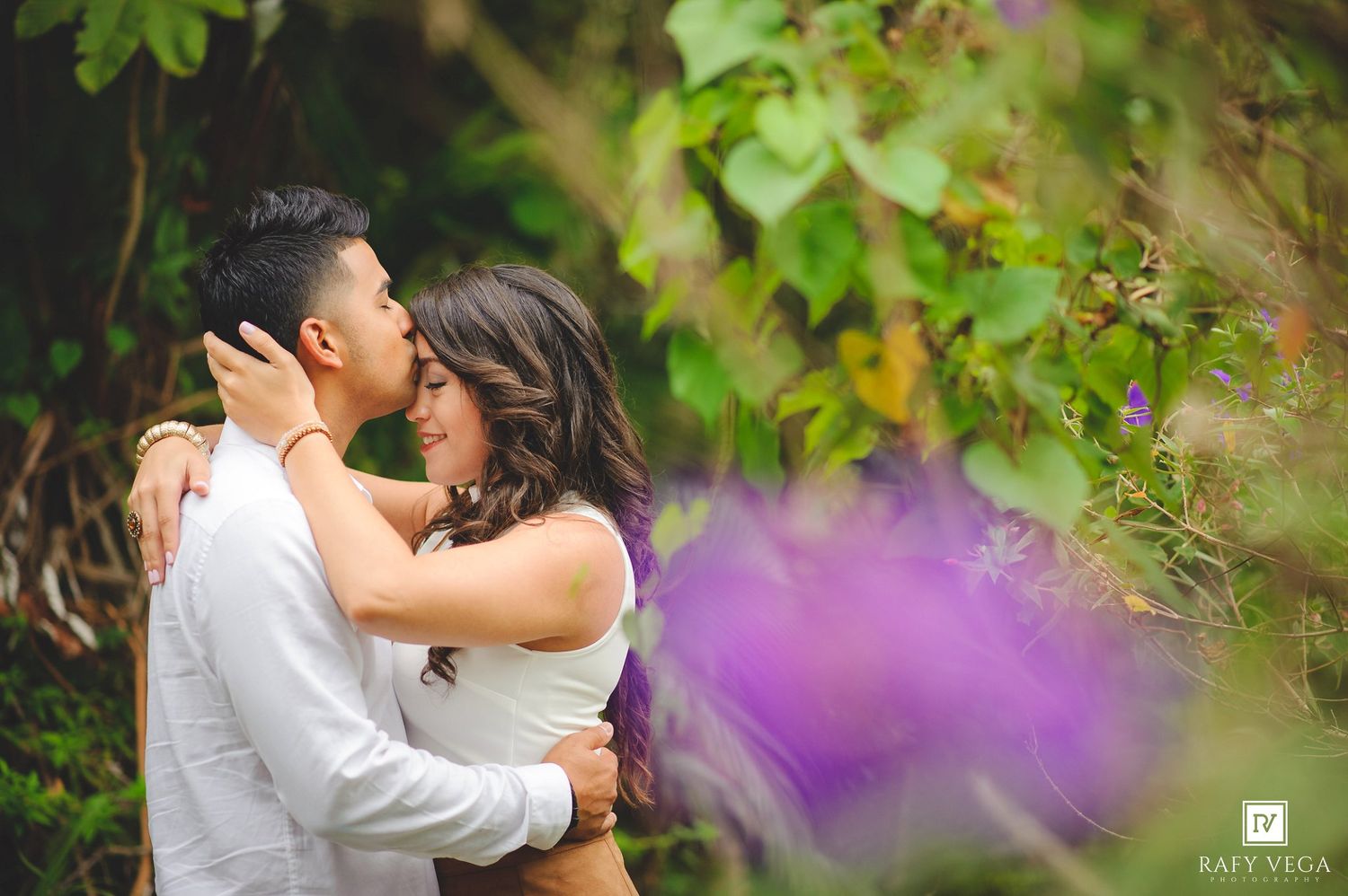 Couple sharing a romantic kiss surrounded by lush green foliage and purple flowers in an outdoor setting.