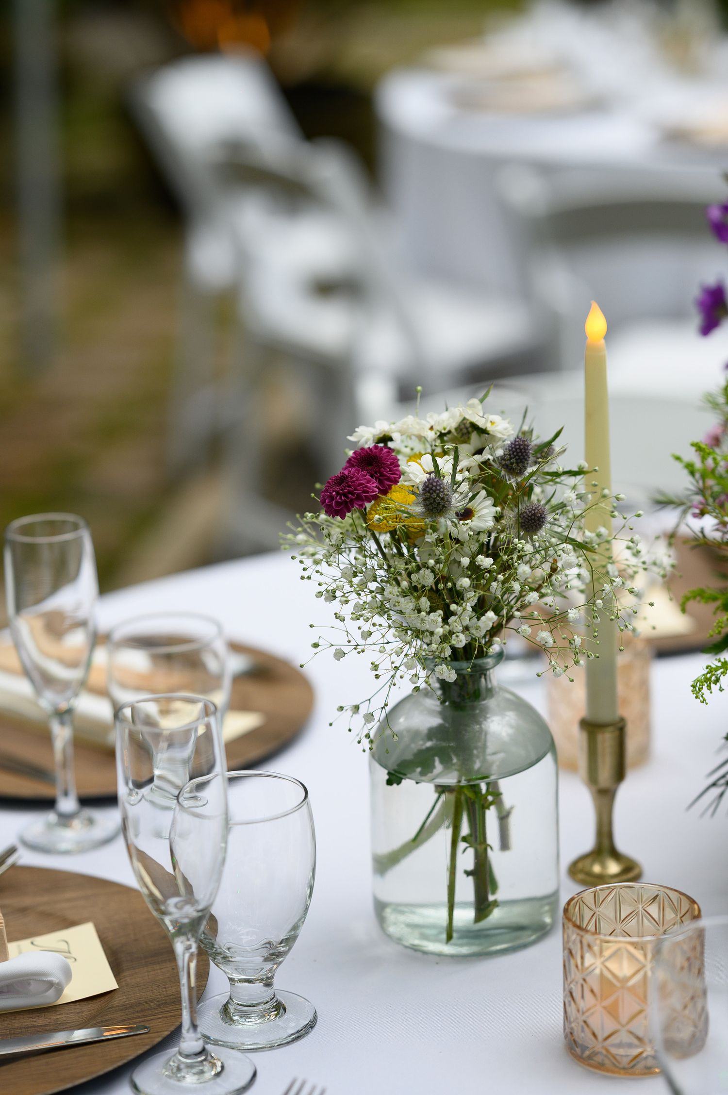 Elegant outdoor wedding reception table with wildflower centerpiece, candle, glassware, and wooden charger plates.
