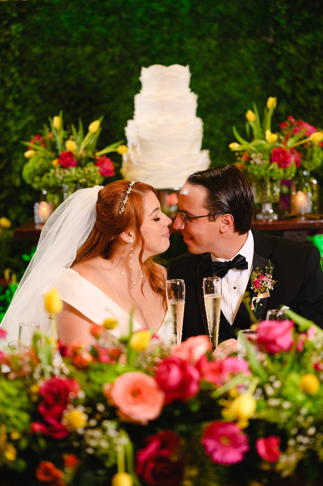 Bride and groom share a kiss at wedding reception with white tiered cake and colorful floral arrangements in background.