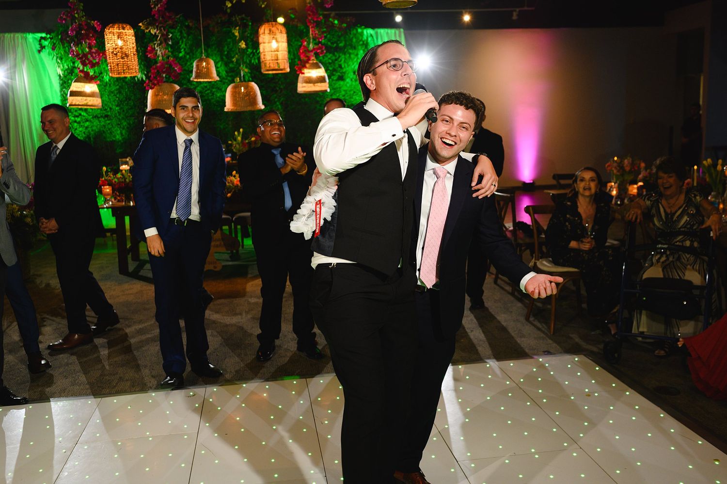 Two men celebrating joyfully on a dance floor at an elegant wedding reception with decorative lighting.