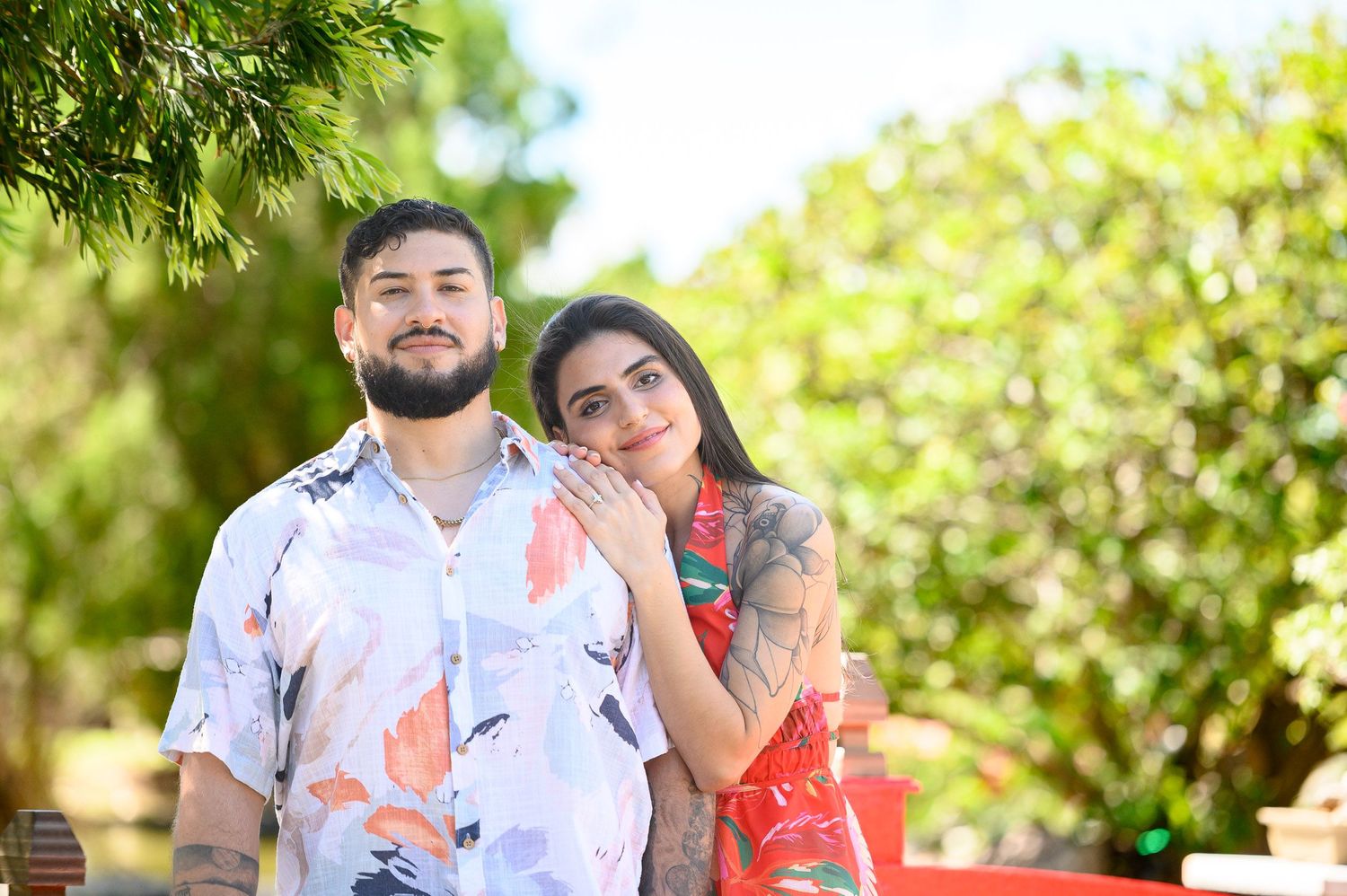 A couple poses outdoors, man in floral shirt and woman in red dress with tattoos, surrounded by lush green trees.