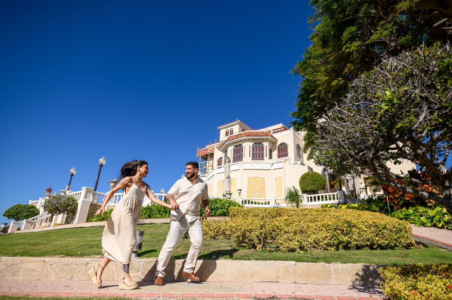 Couple dancing joyfully outside a grand Mediterranean-style mansion with lush green gardens under a bright blue sky.