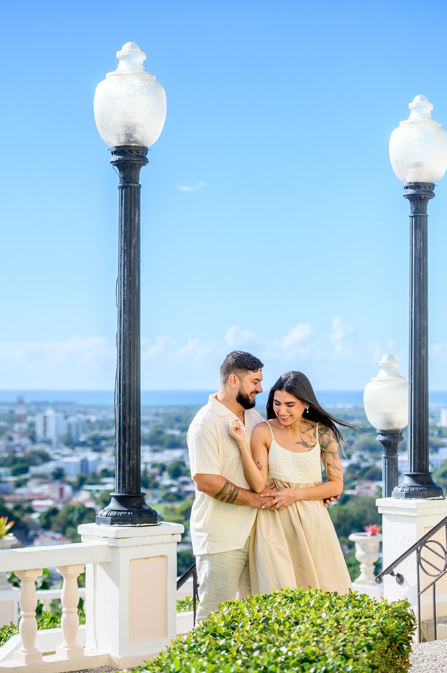 Romantic couple embracing on a scenic overlook with ornate lamp posts and city skyline under a bright blue sky.