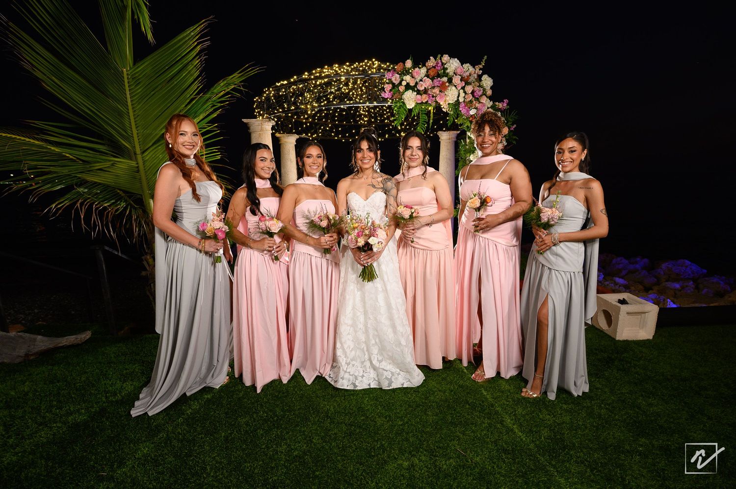 Bride in a white gown stands among six bridesmaids in pink and gray dresses holding bouquets before a floral wedding arch at night.