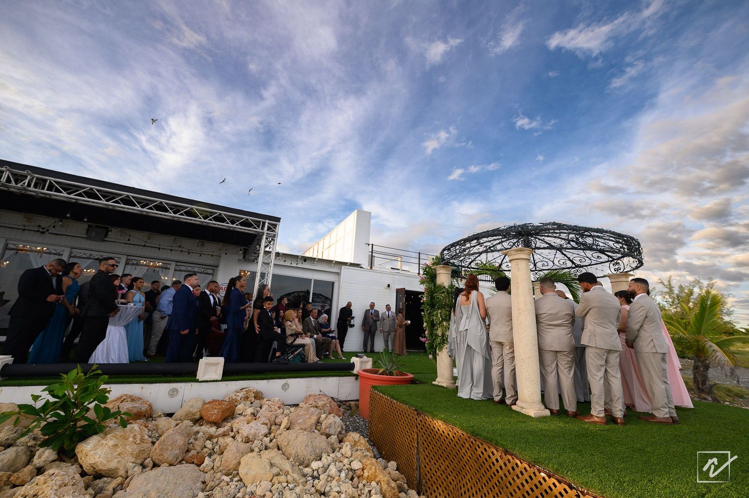 Wedding guests gather on a green lawn beside a circular pergola at an outdoor ceremony venue under a bright blue sky with wispy clouds.