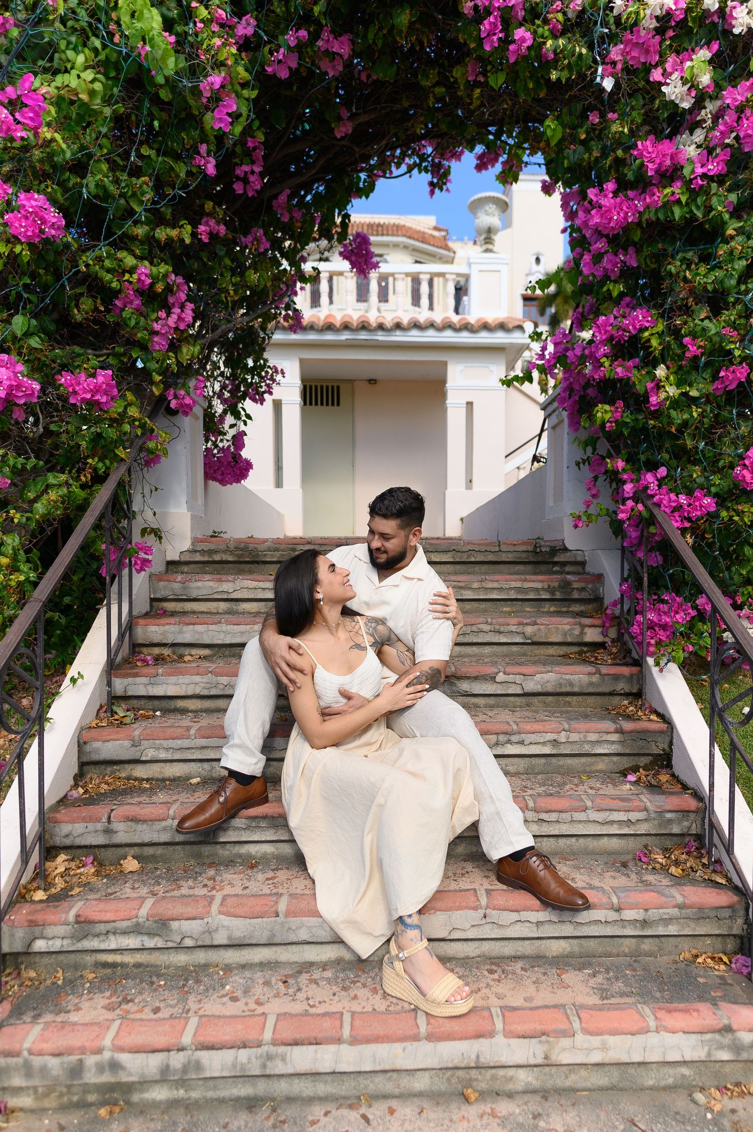 Couple sitting on brick steps under a floral arch of pink bougainvillea, embracing in front of a white villa.