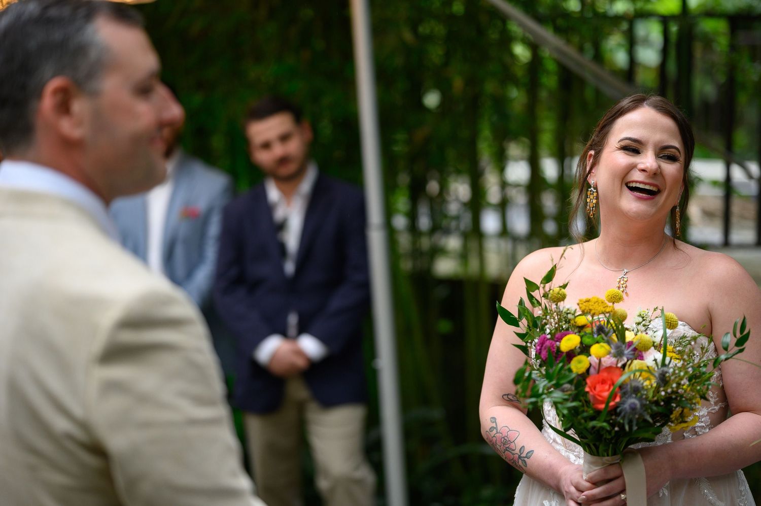 Smiling bride holding colorful wildflower bouquet during outdoor wedding ceremony with groom and groomsman in background.