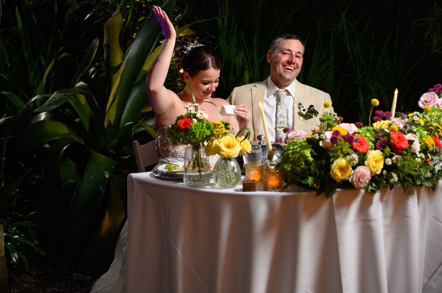 Bride and groom laughing at sweetheart table adorned with colorful floral arrangements at elegant outdoor wedding reception.