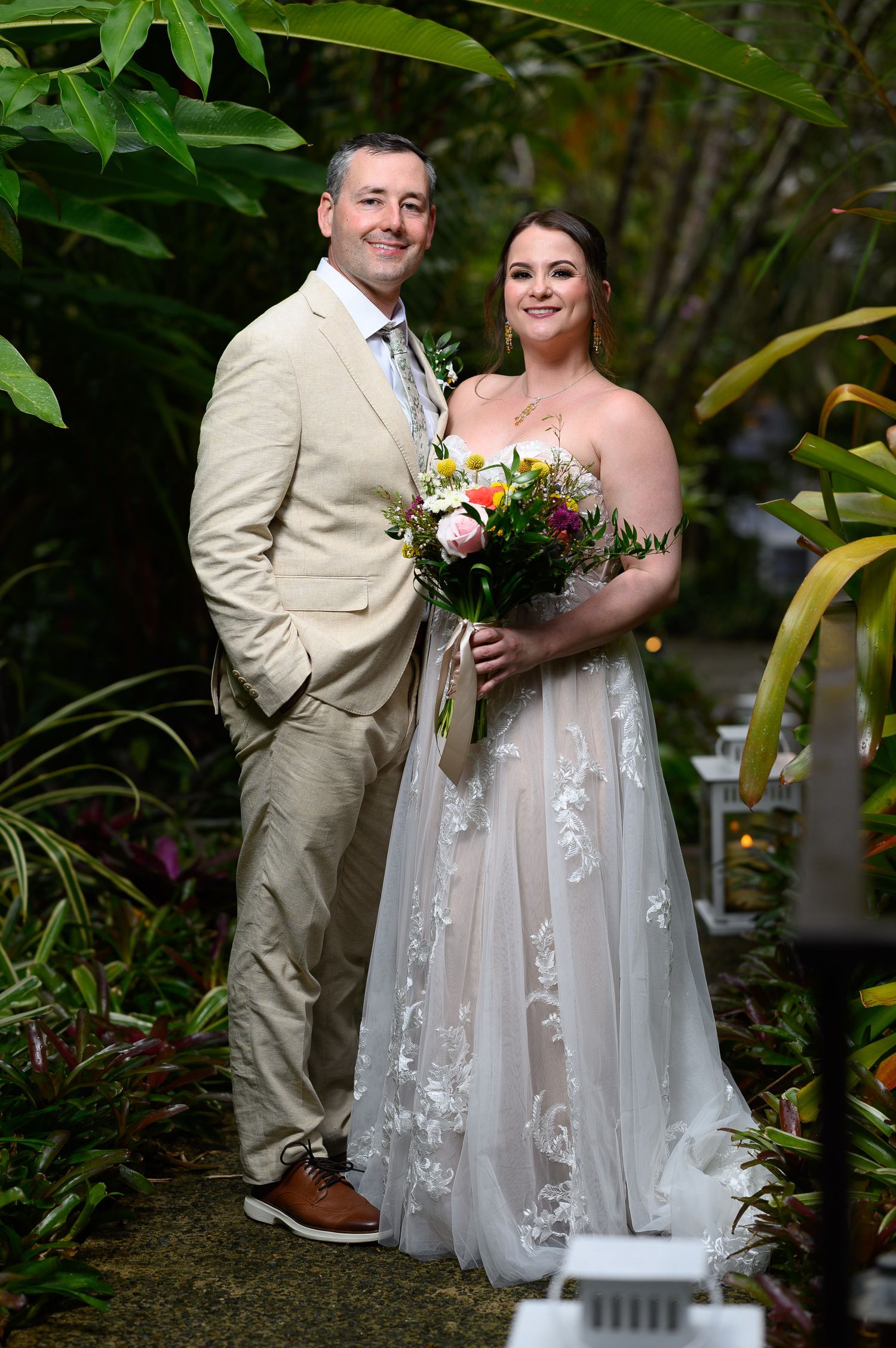 Bride and groom posing together in a lush garden, bride holding a colorful bouquet in a white lace gown.