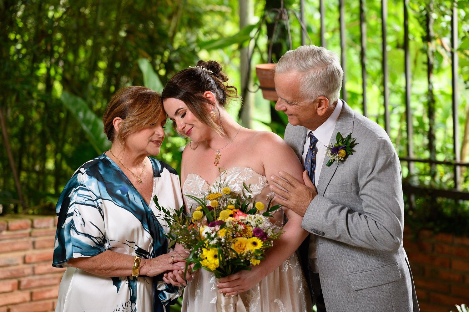 Bride in white gown holds yellow floral bouquet with elderly couple smiling outdoors near green foliage.