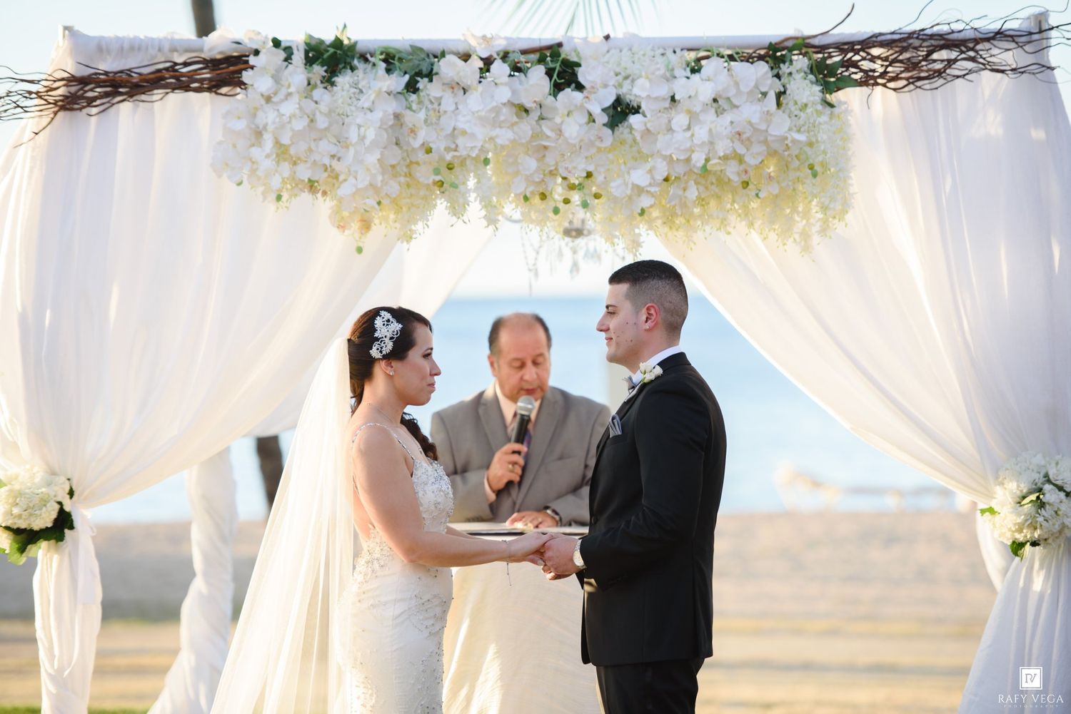 Couple exchanging vows under a white floral arch with draped fabric at an outdoor wedding ceremony.