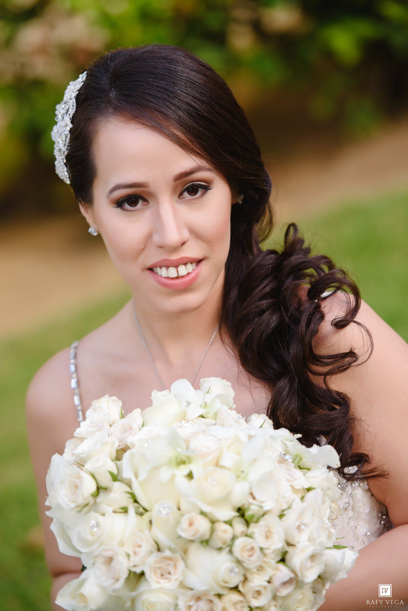 Bride with dark hair holding white rose bouquet, wearing crystal hair accessory and wedding dress outdoors.