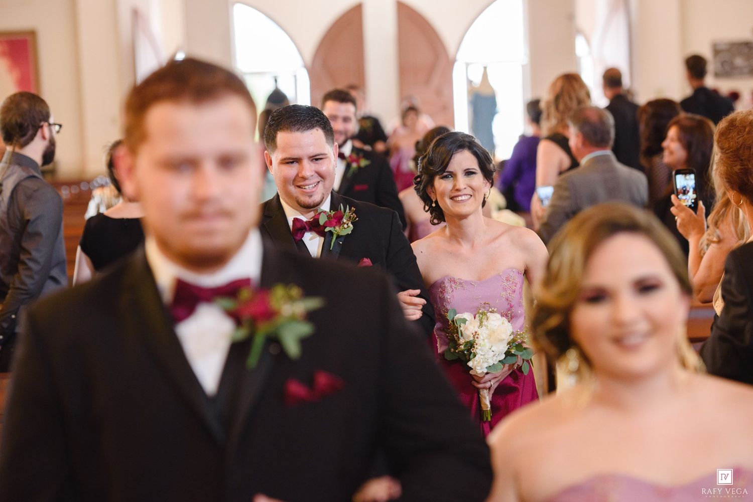 Smiling wedding party walks down church aisle, groomsman escorts bridesmaid in purple dress at reception.