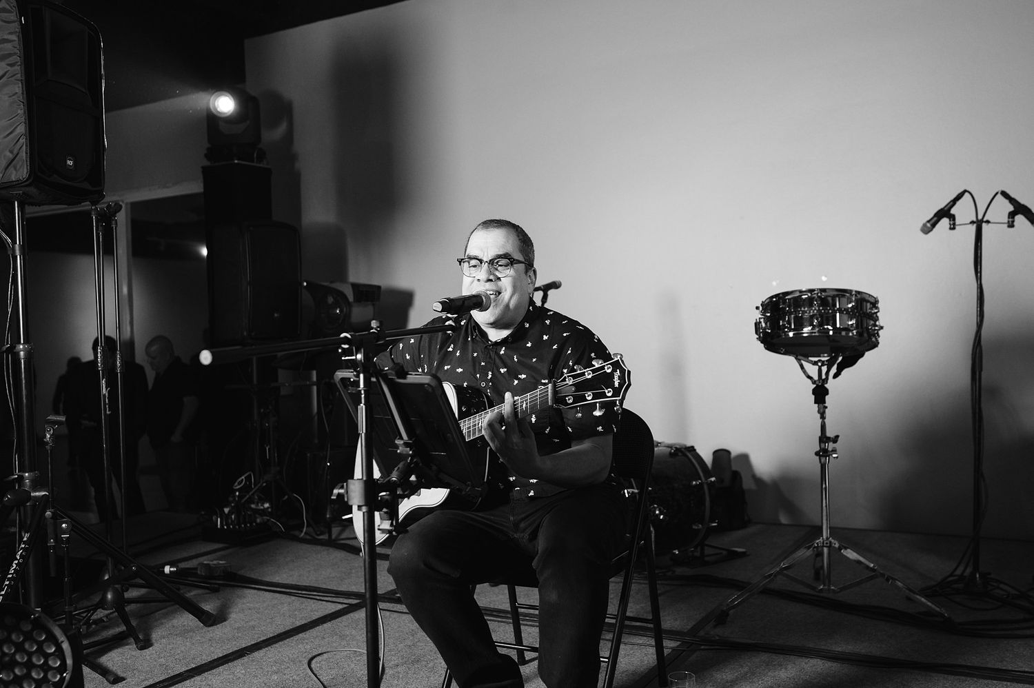 Male musician plays acoustic guitar while seated at microphone stand in black and white studio recording session.