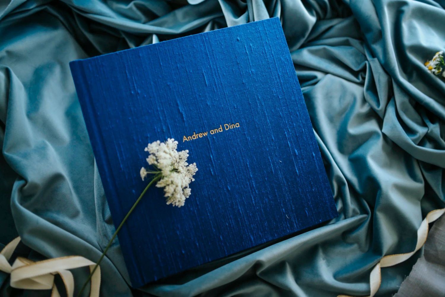 Blue silk wedding photo album with white flowers, personalized with Andrew and Dina names, on teal satin fabric.