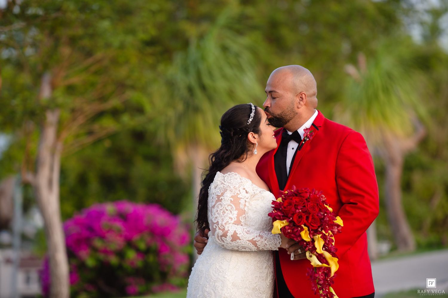 A bride and groom share a romantic moment outdoors, with the groom in a red tuxedo and bride holding a red bouquet.