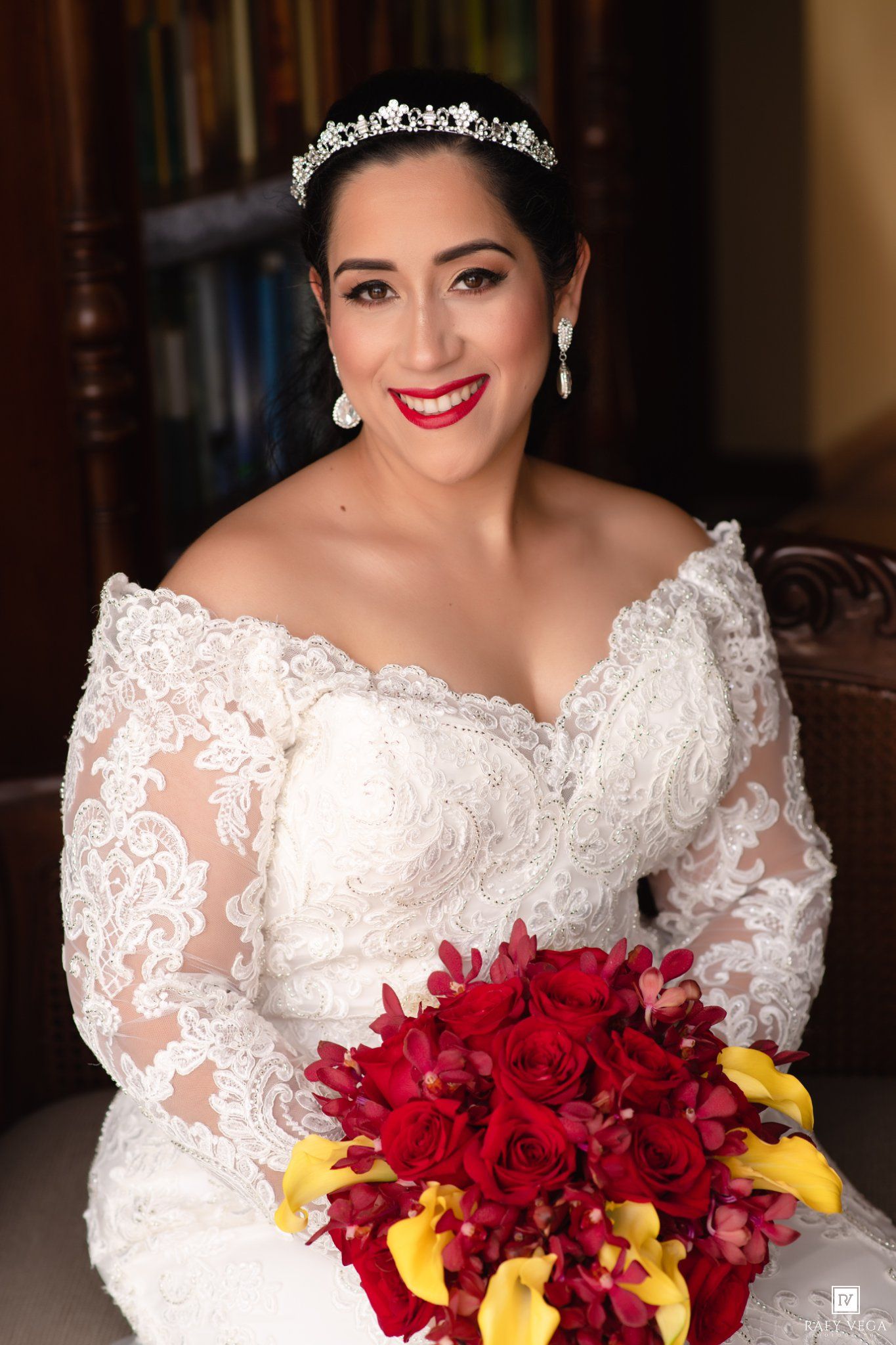 Smiling bride in white lace off-shoulder gown and tiara holds red roses and yellow calla lily bouquet.