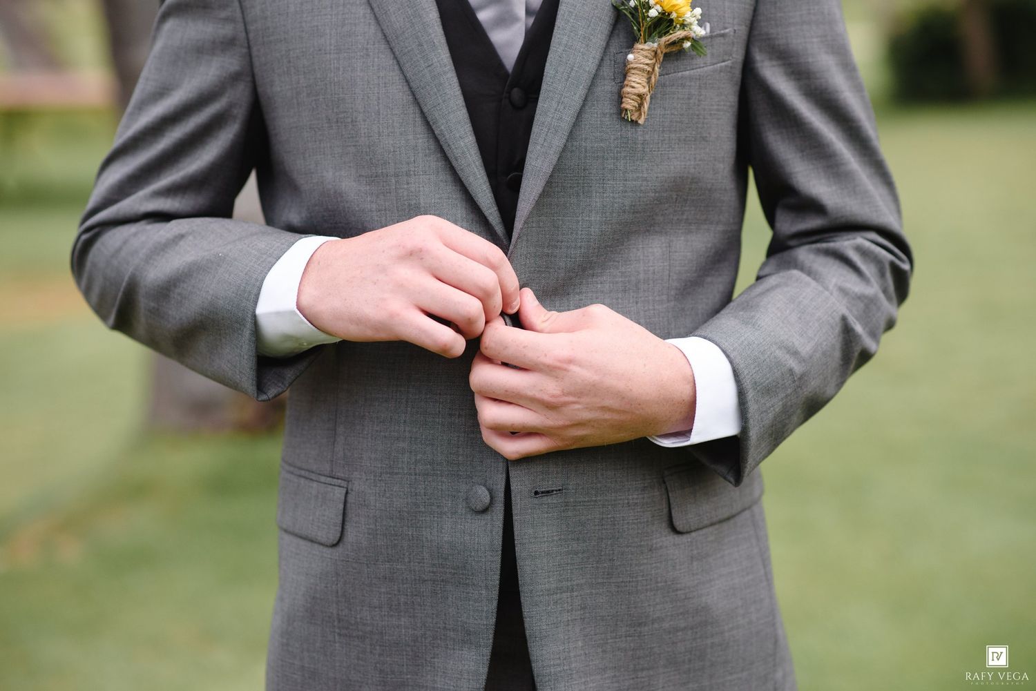 Groom in gray three-piece suit buttoning jacket with floral boutonniere at outdoor wedding ceremony.