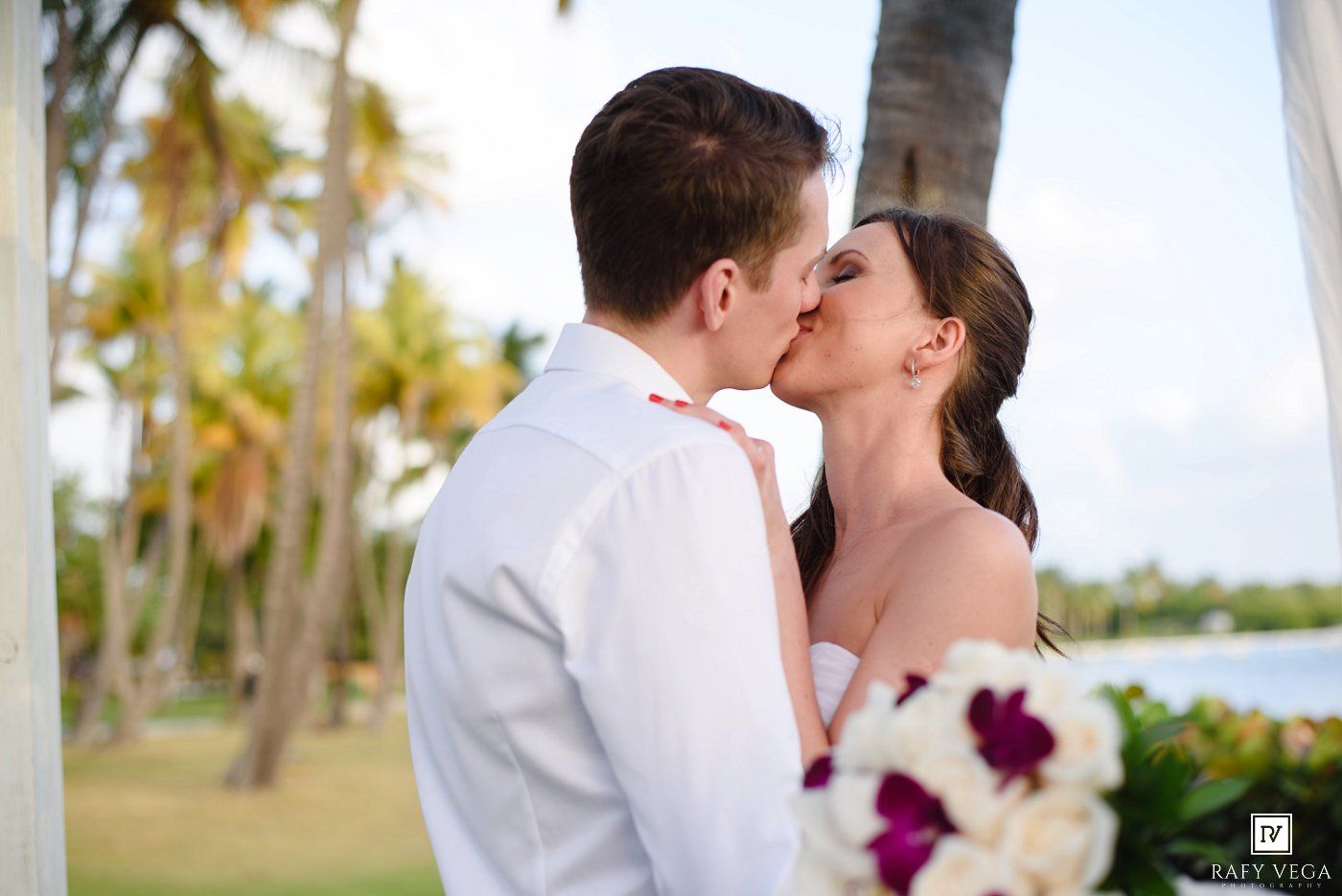 A bride and groom share a romantic kiss at an outdoor tropical wedding ceremony with palm trees in the background.