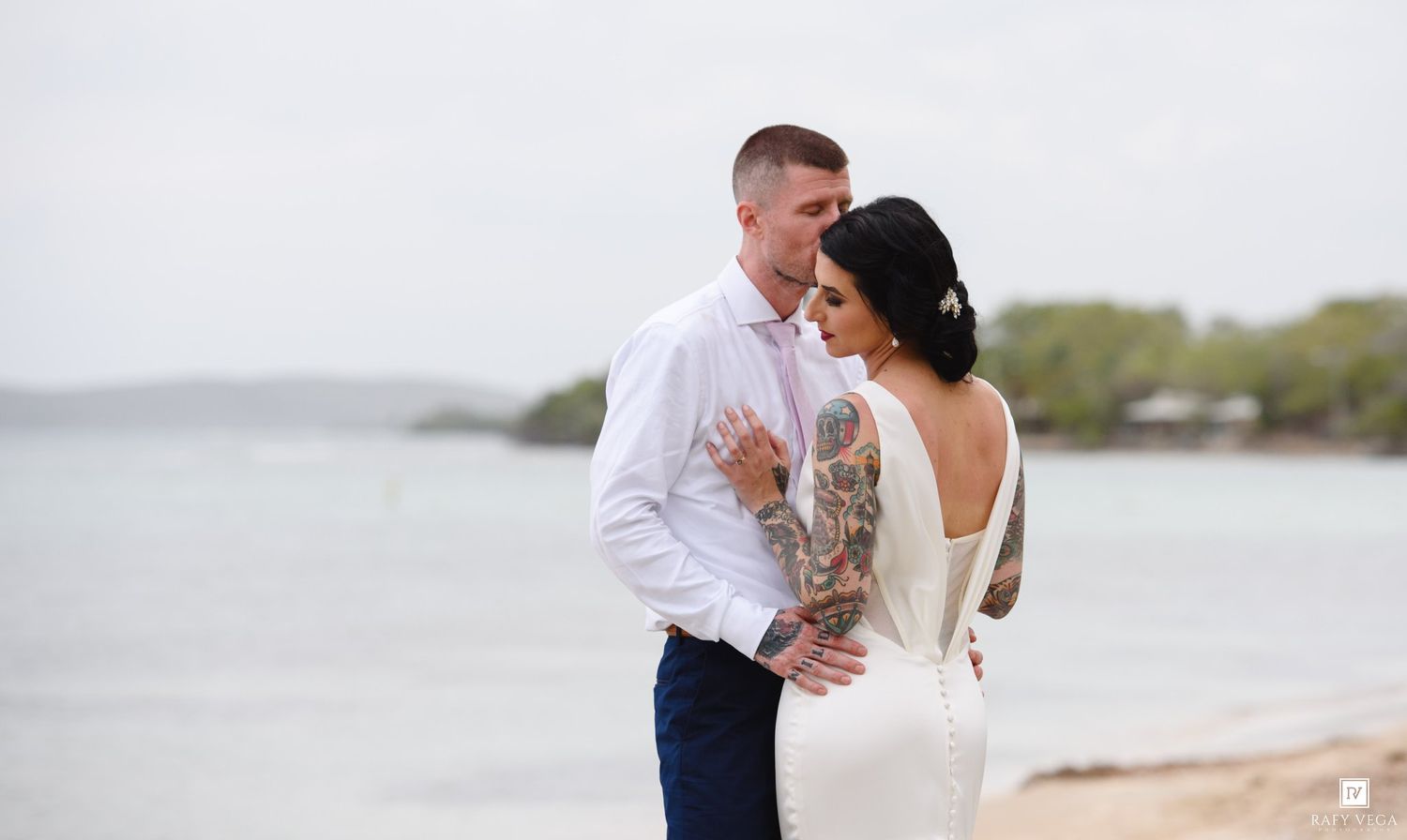 Tattooed groom embraces bride in backless white dress on sandy beach with ocean backdrop.