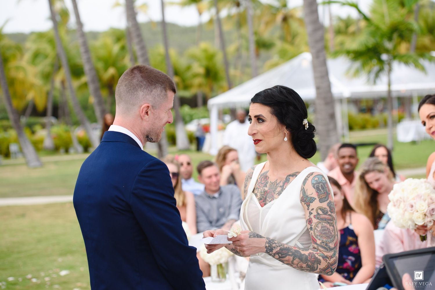 Bride with tattoos exchanges vows with groom in navy suit at outdoor tropical wedding ceremony.